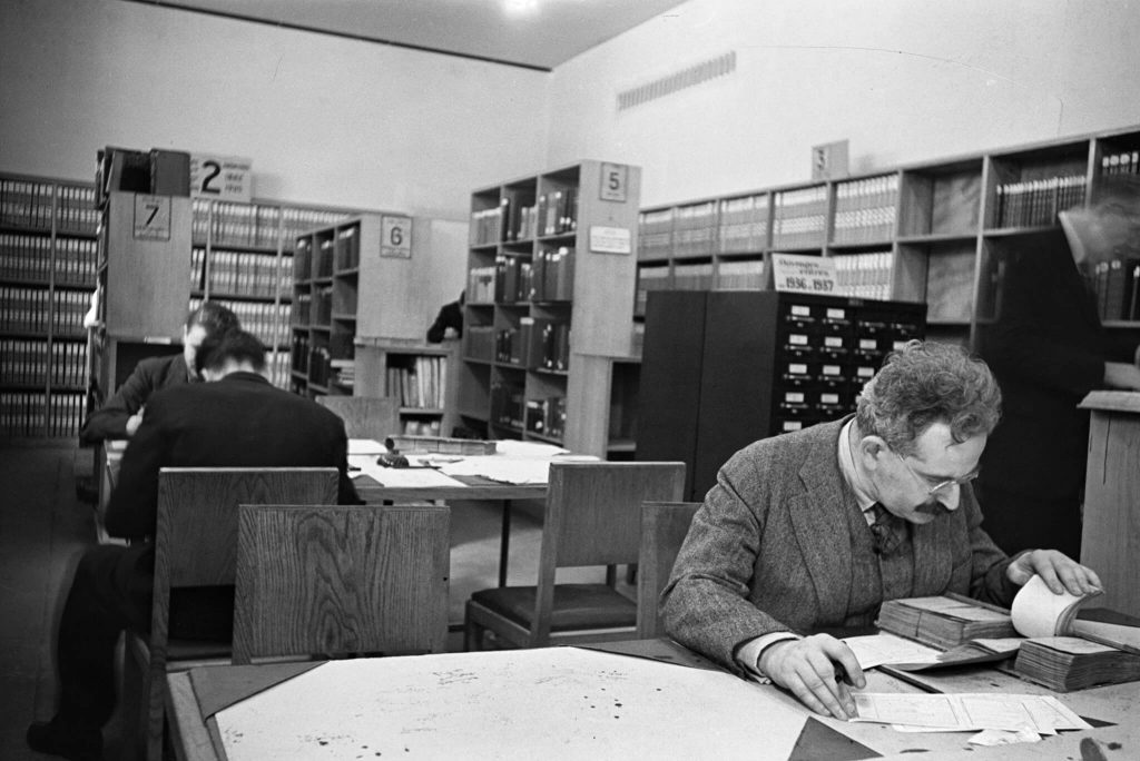 A man at a desk, taking notes from a library catalogue.